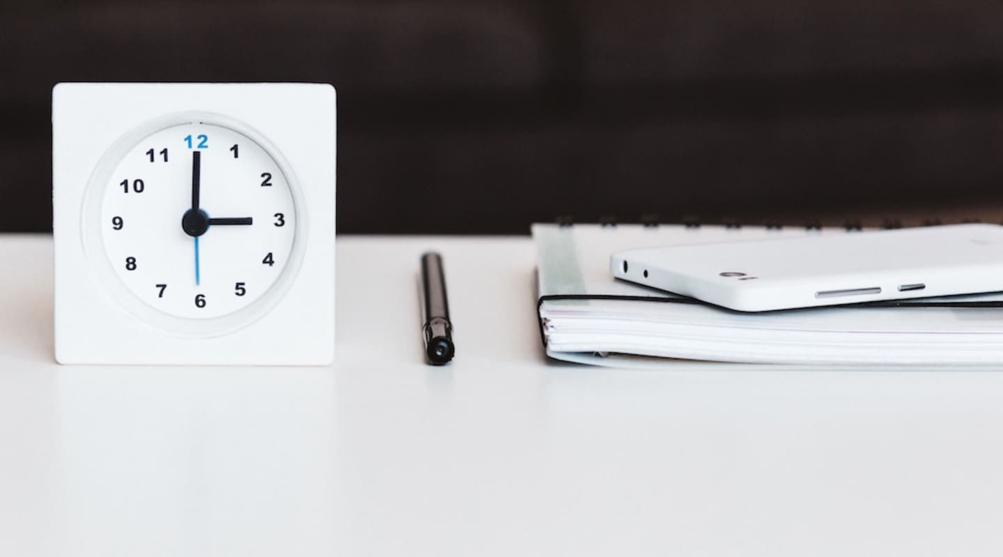 White clock at 3:00, pen, notebook, and phone on white desk. Time management.